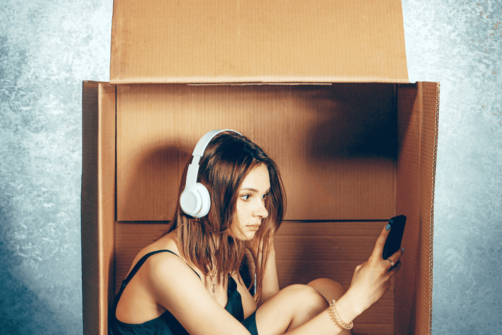 A woman crouched in a large cardboard box with headphone using her phone.Indicating the need for a quiet space or because of procrastination