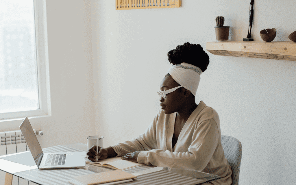Monthly Journaling Prompts. A woman sat at her desk with an open laptop. She is writing in a journal with her planner beside her.