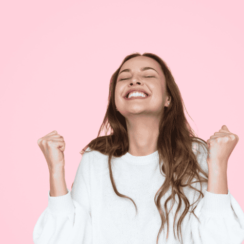 A woman wearing a white jumper has her fists clenched and raised in a mini cheer. Her eyes are closed and she is smiling