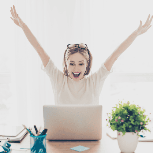 woman sat at desk with arms outstretched cheering. Looking at her laptop on the desk in front of her