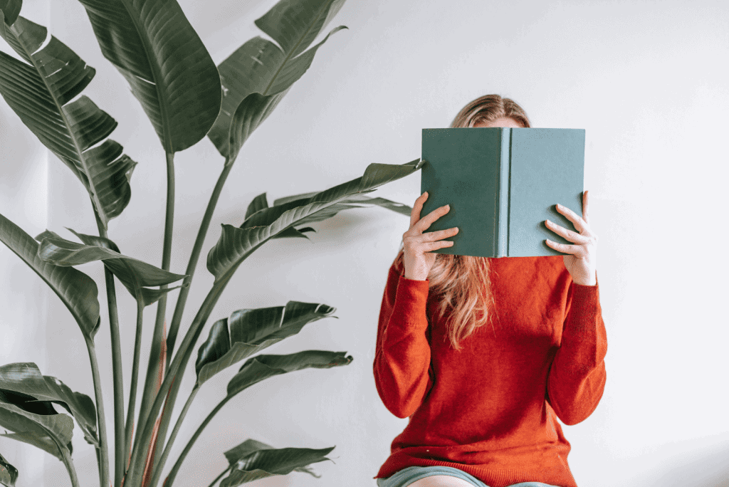 A woman in an orange jumper sat down with a large green plant behind her. She is holding a pale green book in front of her face with the pages open so that it is blocking her face