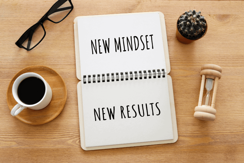 A book opened on a brown desk with the words "NEW MINDSET NEW RESULTS" written. Next to the book are glasses, a cup and small plant. Encouraging Self confidence