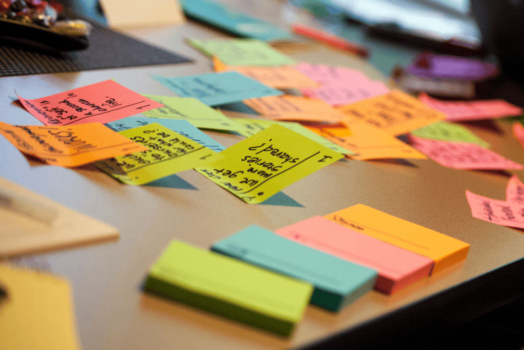 A desk with multiple post it notes spread around with writing on them. Then a pile of 4 different coloured post it note pads without writing yet. It depicts a busy schedule where time management may be impacted