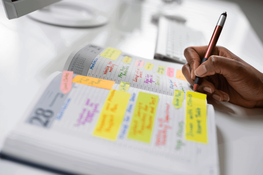 Image of someone writing in a planner. The planner has lots of writing and colourful labels inside it. Time management planner