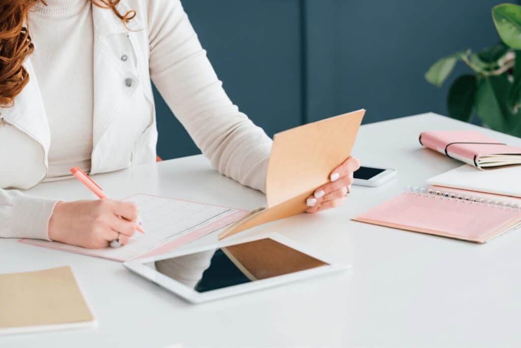 Passive income. A women wearing a white jacket sat at a desk holding a notebook and pen for a tablet. Other books on the white desk around her.