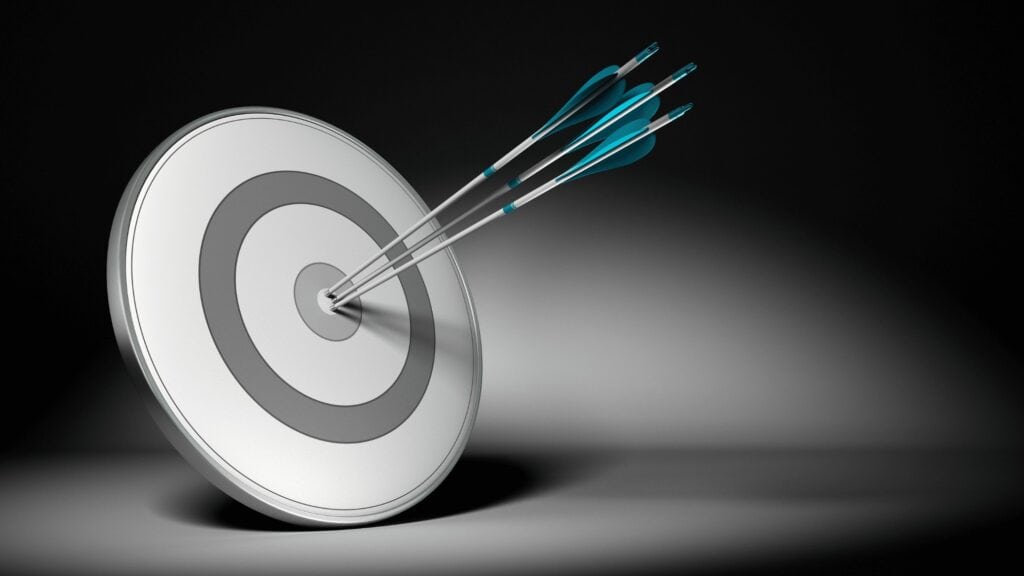A black shadowy background with a white target board with two blue arrows in. It is used to indicate hitting the mark in your coaching business with you product suite