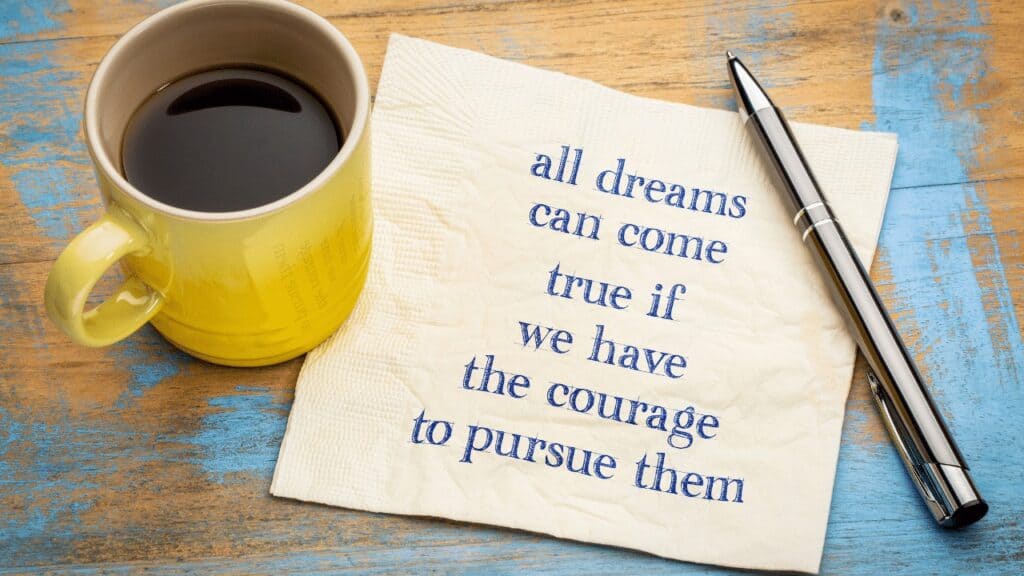 Achieving goals motivation - An image of a yellow mug with black coffee on a blue stained wooden table with a piece of paper saying "All dreams can come true if we have the courage to pursue them"