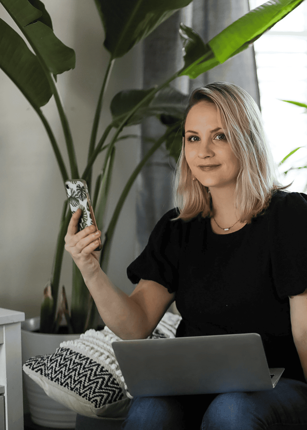 An image of a lady with blond hair, wearing black t shirt. Sitting down. Large plant behind her. Laptop on knees & holding a phone,