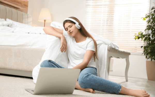 A woman sat on a sofa with headphones on looking at her open laptop.