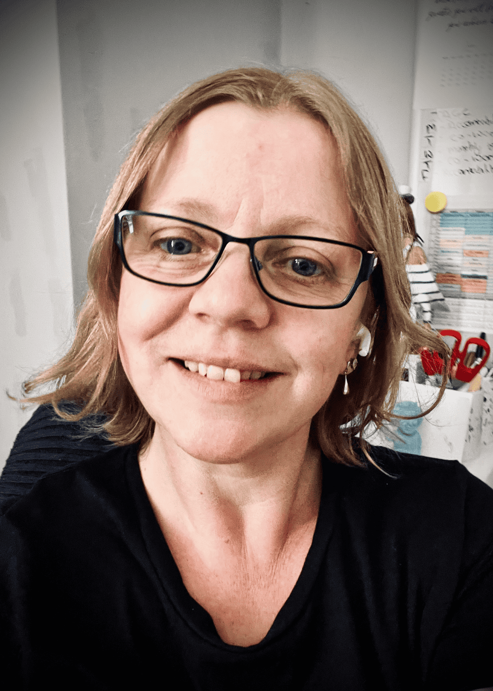 Lynsey Wall, Coach & Mentor at Coach Writes. About me - A headshot of Lynsey smiling. Wearing a black t-shirt, blue framed glasses. Office walls behind her.