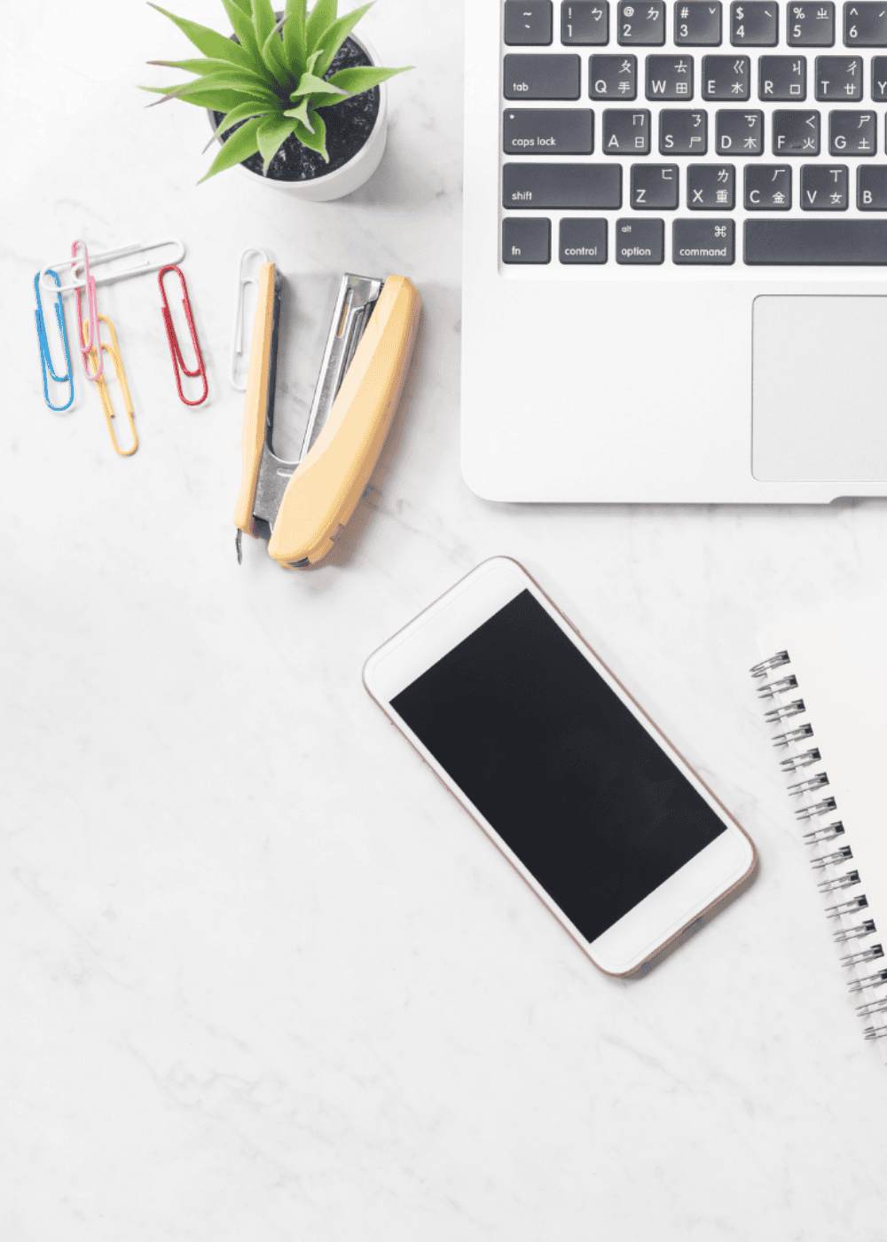 An image of a white desk with a laptop, pens, a phone & stationery.