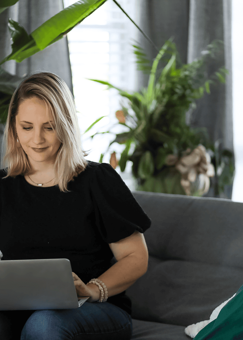 An image of a lady wearing blue jeans & a black t shirt sat at home on a sofa with green cushions. On her lap is a laptop & in the background are plants